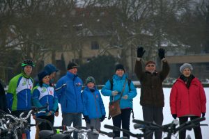 Zdjęcie ośmiu osób tworzących lewą górną część ustawionego na Jasnych błoniach WOŚP-owego serca . W dole widać kierownice i siodełka rowerów które tworzyły dolny zarys serca a nad nimi w centralnej części zdjęcia , uśmiechnięte postacie osób w różnokolorowych strojach . Część osób jest ubrana w stroje rowerowe i ma na głowach kaski rowerowe , inne osoby są w ubraniach wyjściowych z szalikami i w różnokolorowych czapkach. Przekrój wiekowy od 10 do 70 lat . Jedna z osób ma uniesione do góry ręce w geście pozdrowienia . Dolną część tła tworzy biały śnieg a górną zielono- szary , rozmyty , zarys platanów i stojącego za nimi budynku.