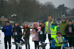 Zdjęcie dziesięciu osób tworzących górną część ustawionego na Jasnych błoniach WOŚP-owego serca . W dole widać kierownice rowerów które tworzyły dolny zarys serca a nad nimi w centralnej części zdjęcia , uśmiechnięte postacie osób w różnokolorowych strojach . Trzy osoby są ubrane w kamizelki odblaskowe NNT w tym jedna w kasku rowerowym i czerwonych okularach , inne osoby są w ubraniach wyjściowych z szalikami i w różnokolorowych czapkach. W centralnej części zdjęcia widać cztery młode wolontariuszki WOŚP z zawieszonymi ma szyjach identyfikatorami i z puszkami w rękach a obok nich , po prawej stronie , pilota akcji NNT uśmiechającego się do fotografa oraz pilotkę akcji NNT stojącą tyłem prezentującą logo projektu NNT . Dolną część tła tworzy biały śnieg a górną parking samochodowy nad którym widać rozmazane drzewa.